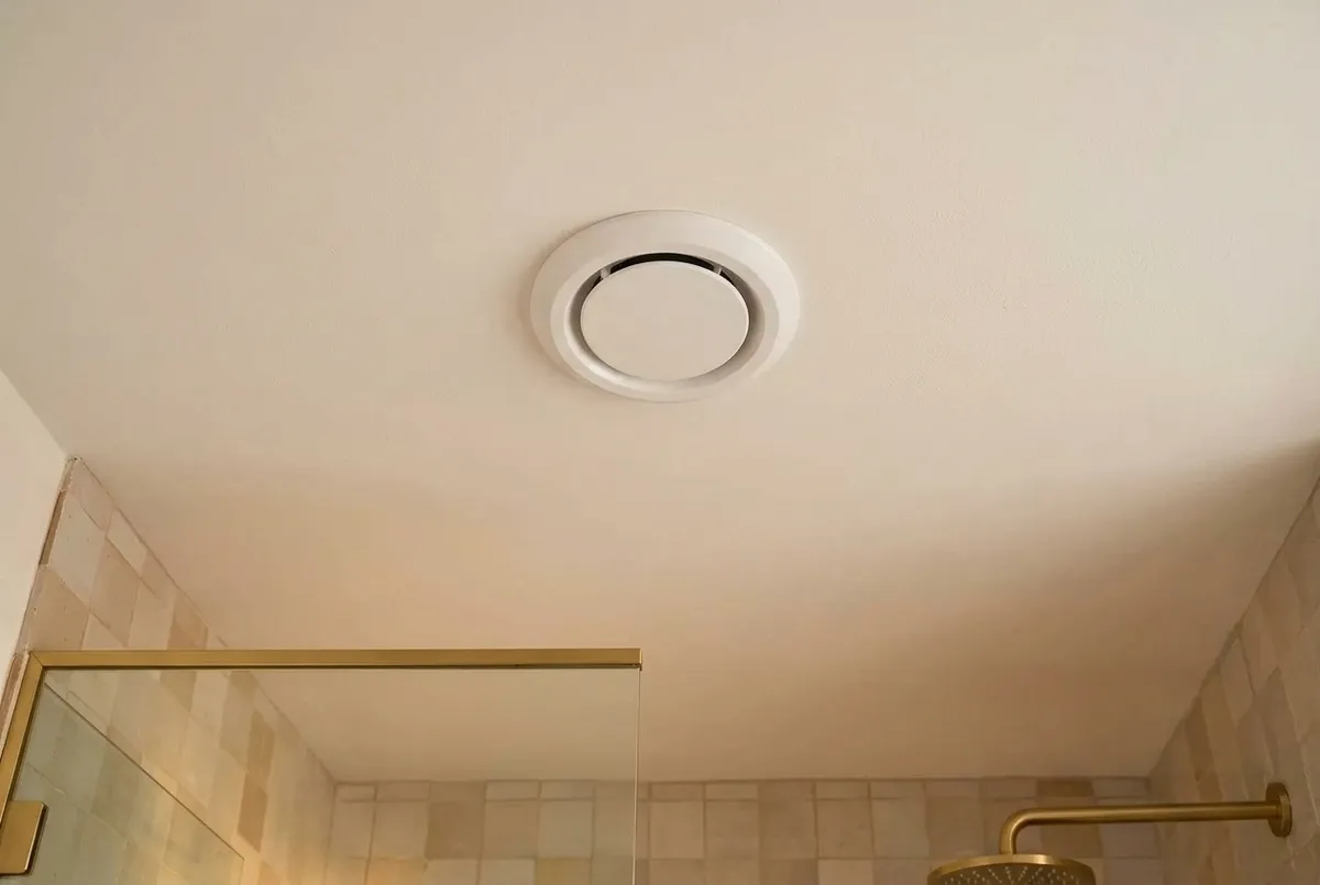 AI-impressie van een rond wit afzuigventiel in een wit badkamerplafond, met onderin beeld de bovenkant van een glazen douchewand en een messing regendouche