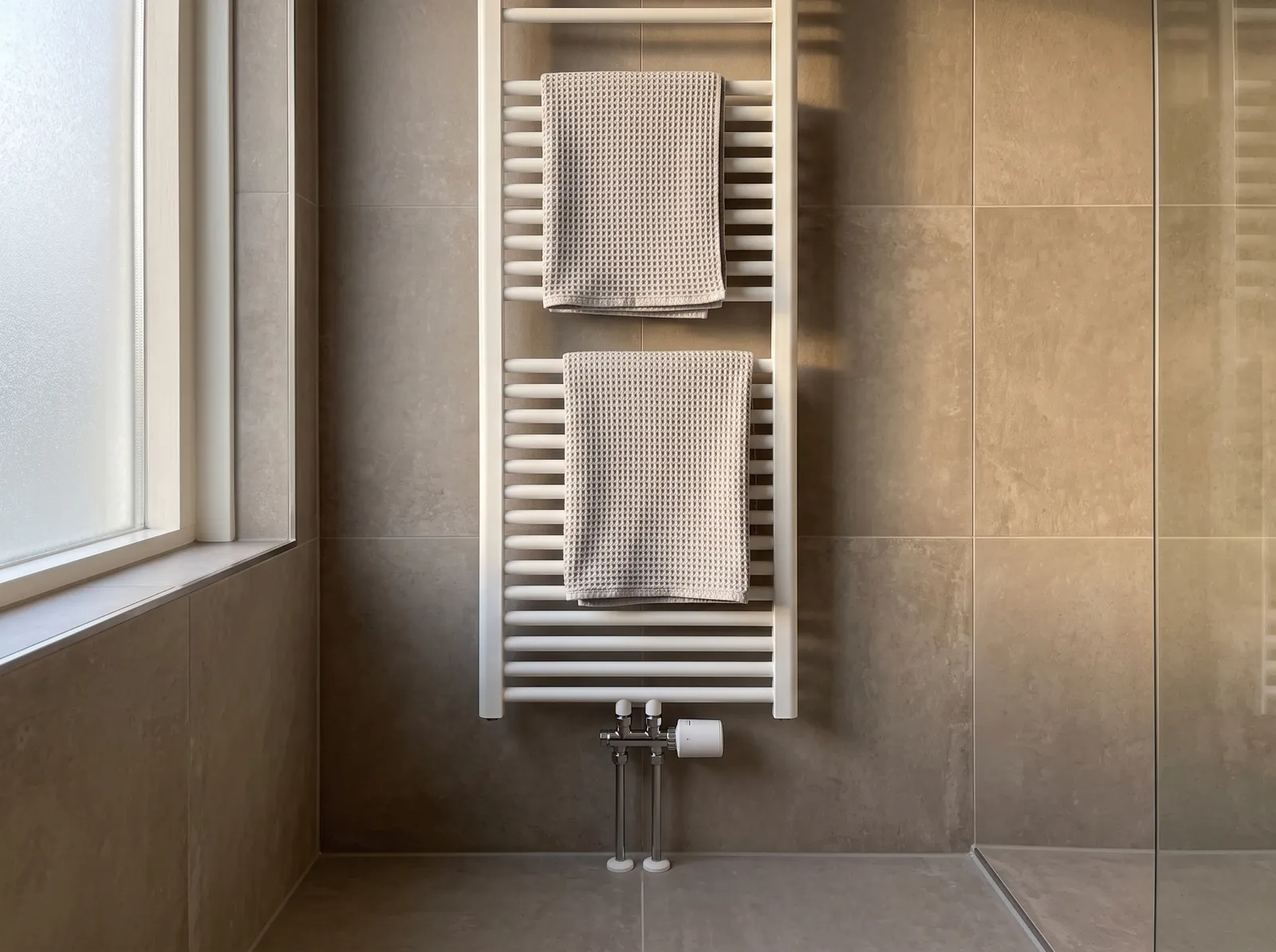AI-impressie van een witte handdoekradiator met twee wafelhanddoeken tegen een wand van grote grijsbeige tegels, met onderaan op het radiatorventiel een witte slimme radiatorknop en ernaast een raam van matglas