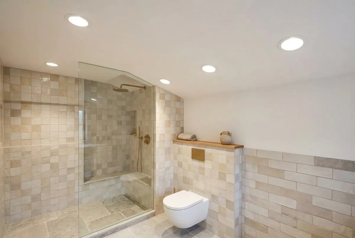AI-impressie van een badkamerplafond met vijf brandende ronde inbouwspots boven een inloopdouche met glazen wand, zandkleurige wandtegels en een hangtoilet