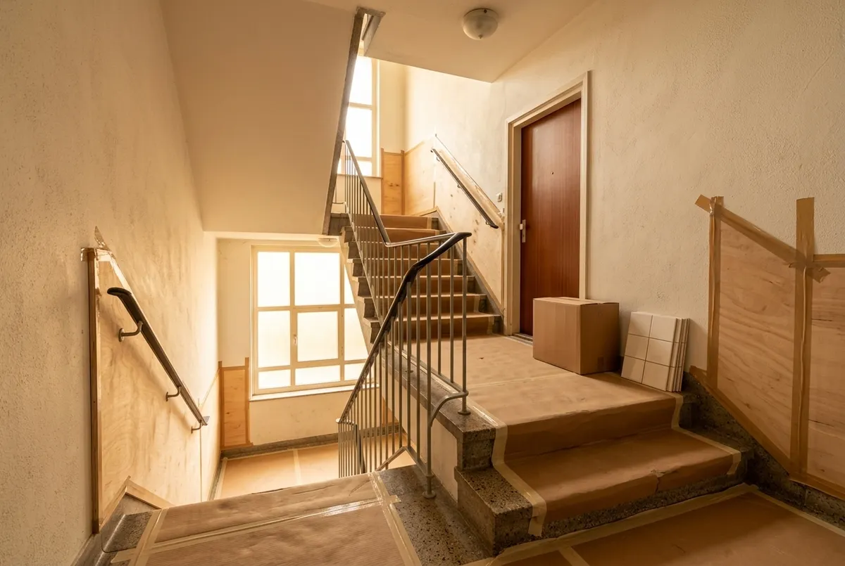 AI-impressie van een trappenhuis in een appartementengebouw met karton op de traptreden en het bordes en houten platen tegen de muren, met een doos en een paar wandtegels naast een voordeur