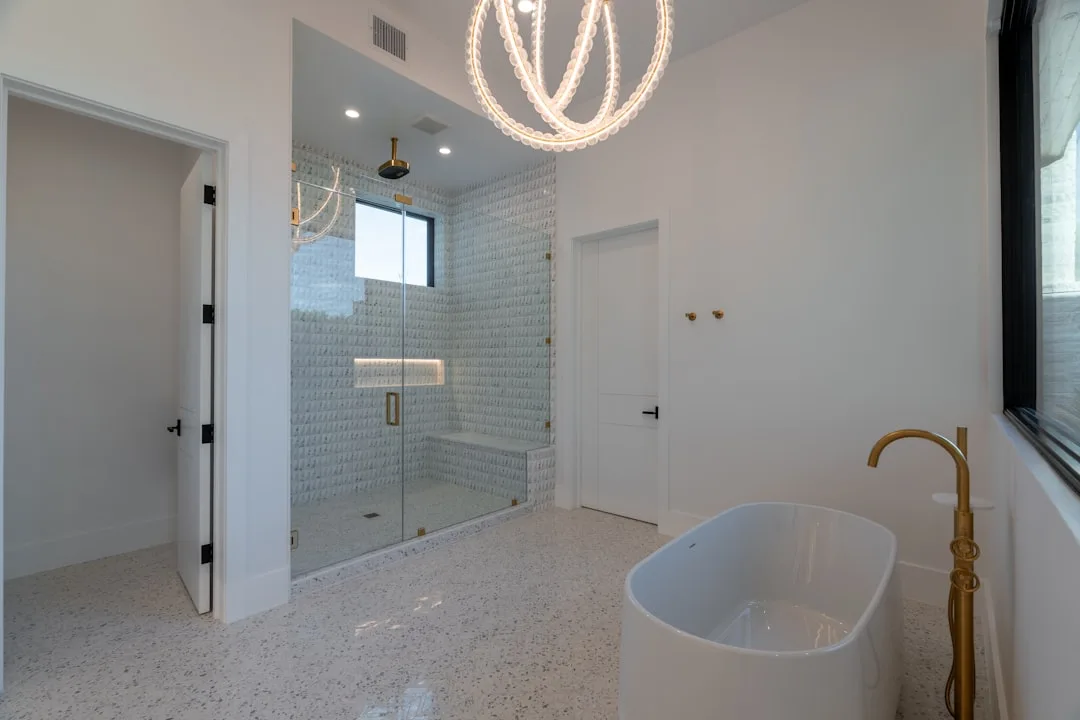 Witte badkamer met terrazzovloer en een vrijstaand bad, naast een betegelde doucheruimte met mozaïektegels, een ingebouwde zitbank en een scharnierende glazen deur
