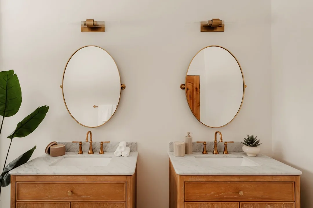 Twee houten wastafelmeubels naast elkaar met marmeren bladen en ovale spiegels in een gouden lijst