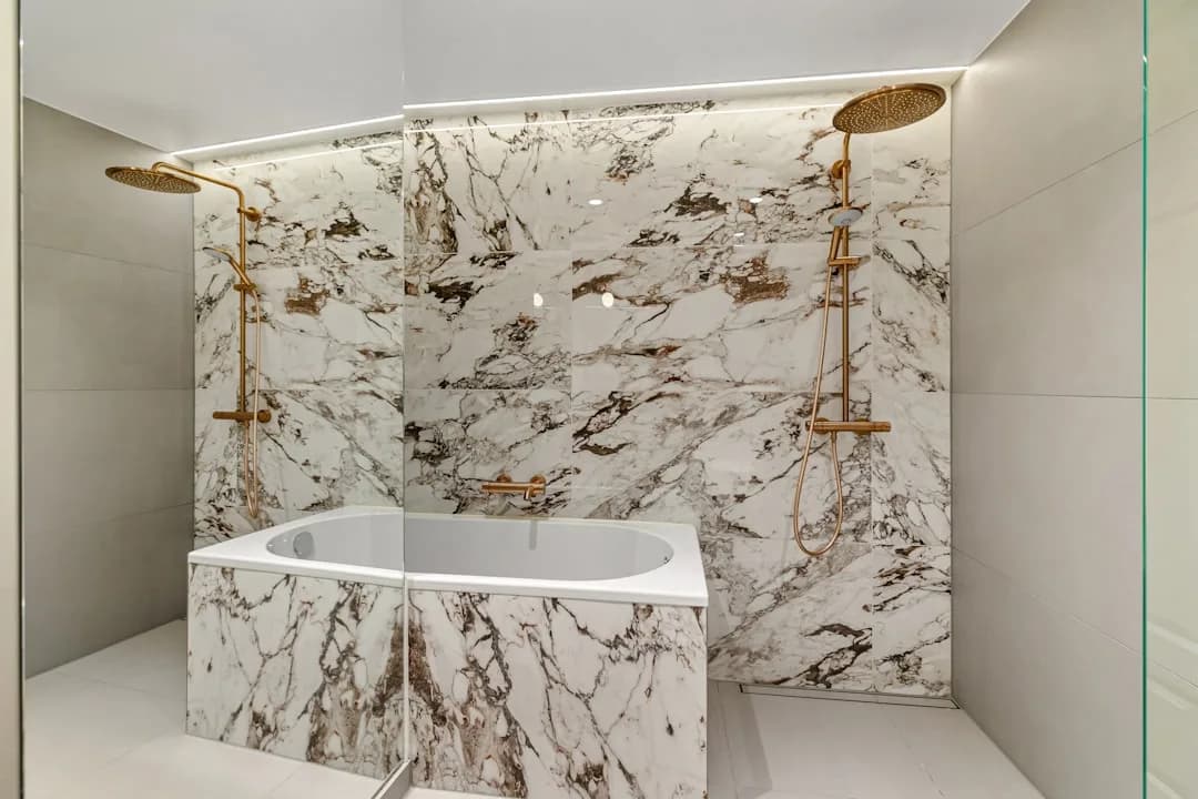 Badkamer met wit geaderd marmer op de wanden, een ingebouwd bad en een inloopdouche met gouden doucheset