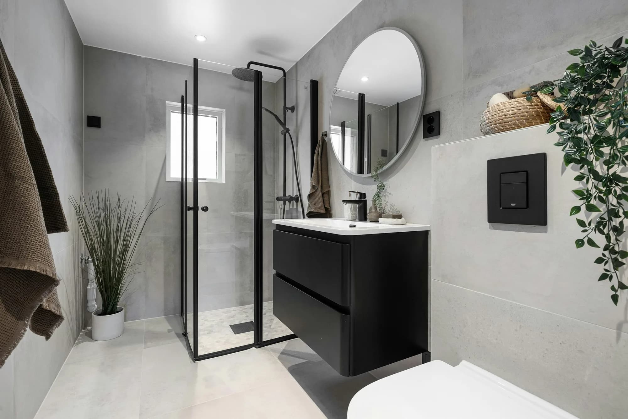 Kleine badkamer met grijze tegels, glazen douchewand, zwart badmeubel en ronde spiegel
