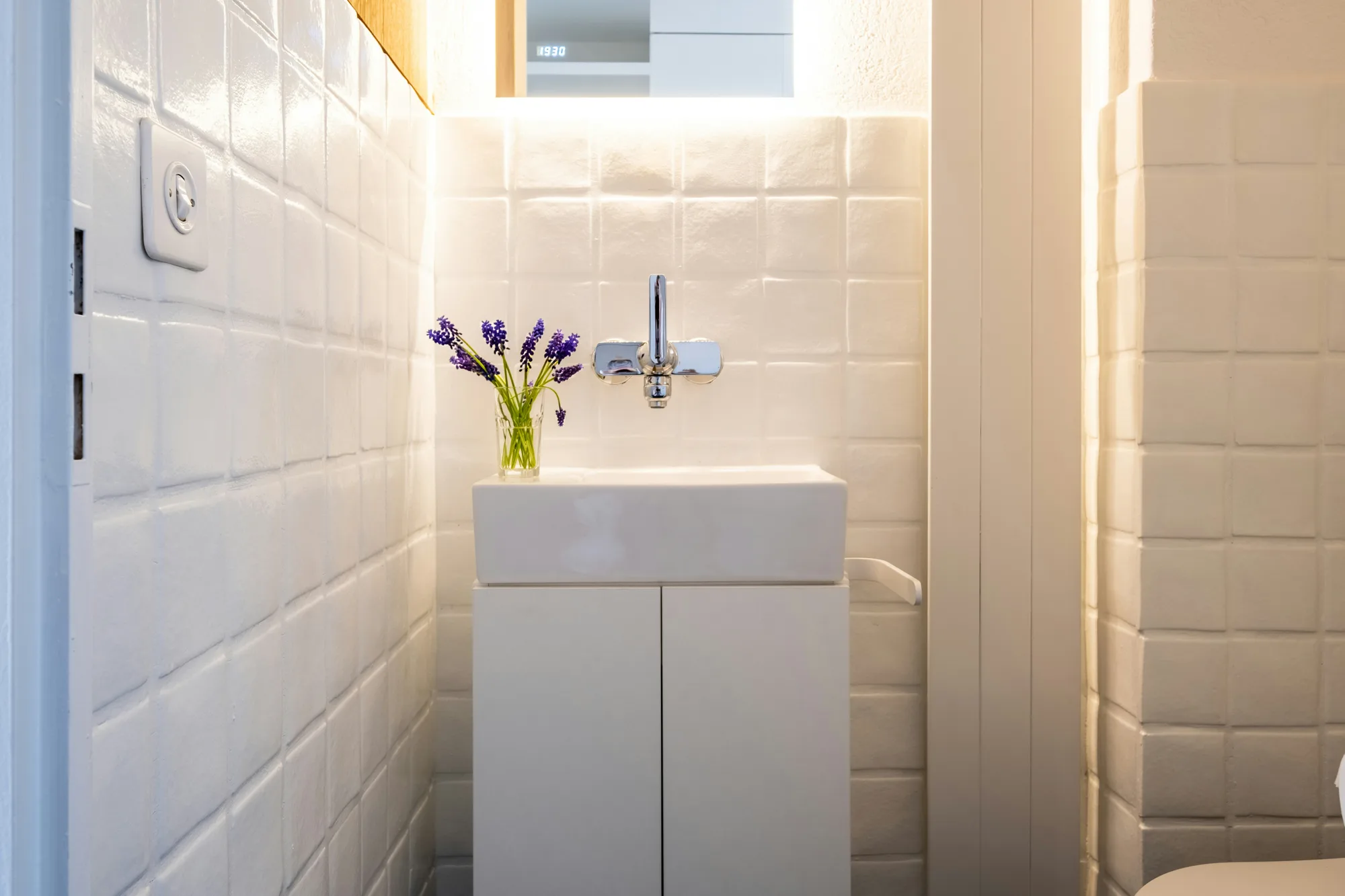 Smalle toiletruimte met witte tegels, een fonteintje met kastje en een vaasje lavendel