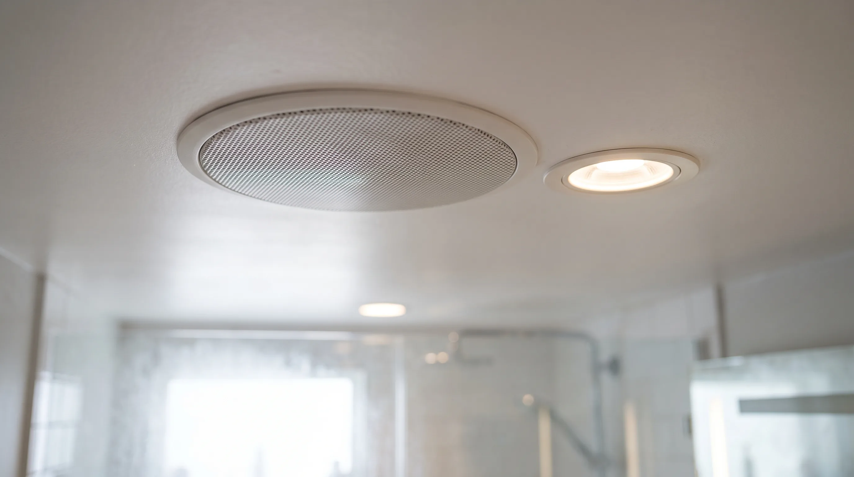 Inbouwspeaker naast een spot in een wit badkamerplafond