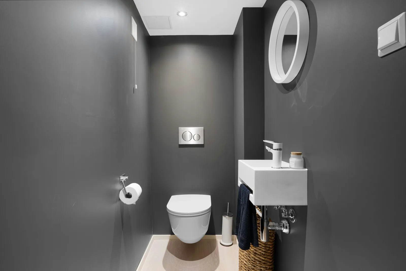 Smalle donkergrijze toiletruimte met een hangtoilet, een klein wandfonteintje en een ronde spiegel