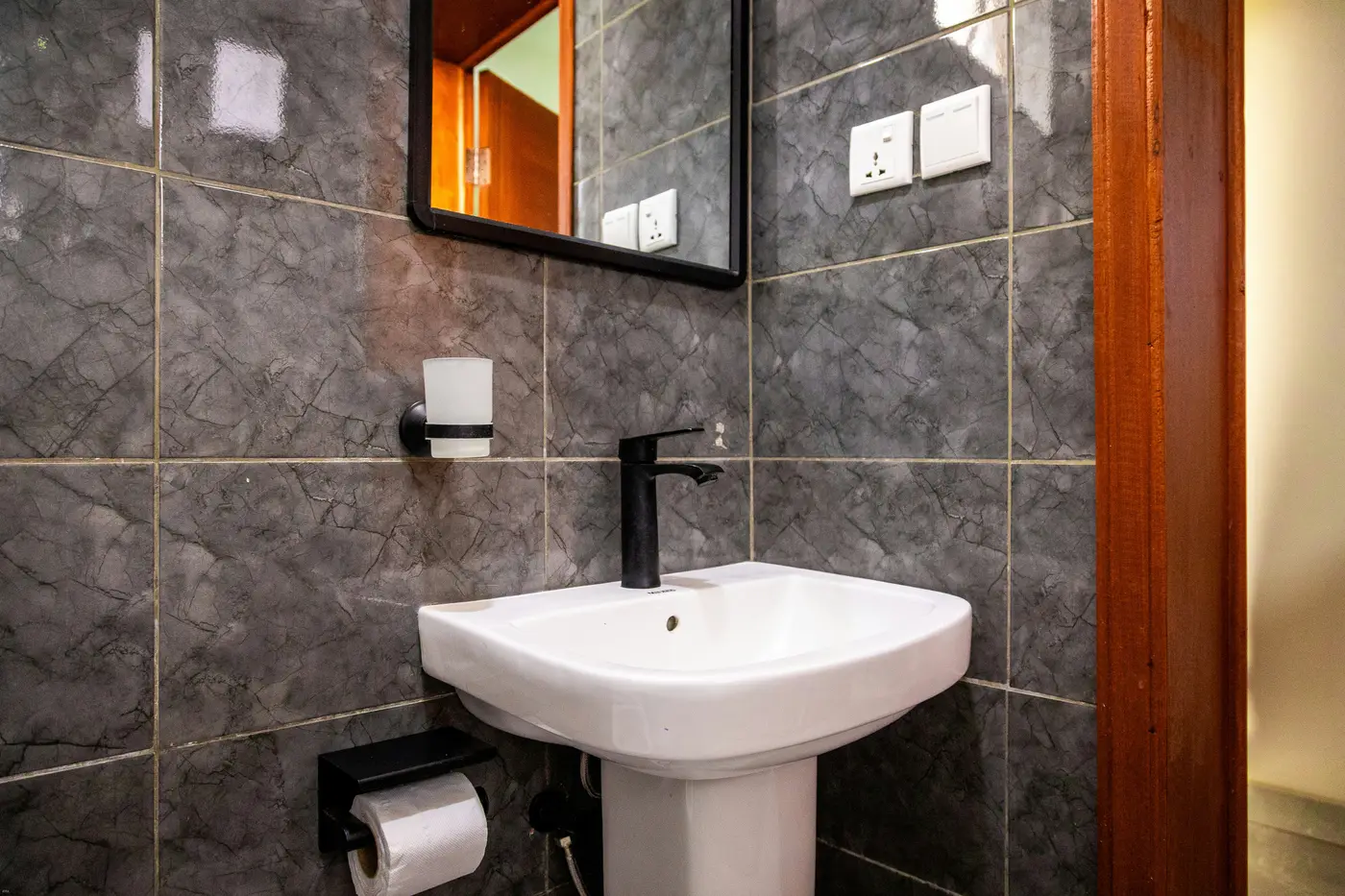Grijs betegelde toiletruimte met een wastafel op zuil, een zwarte kraan en een spiegel met zwarte lijst