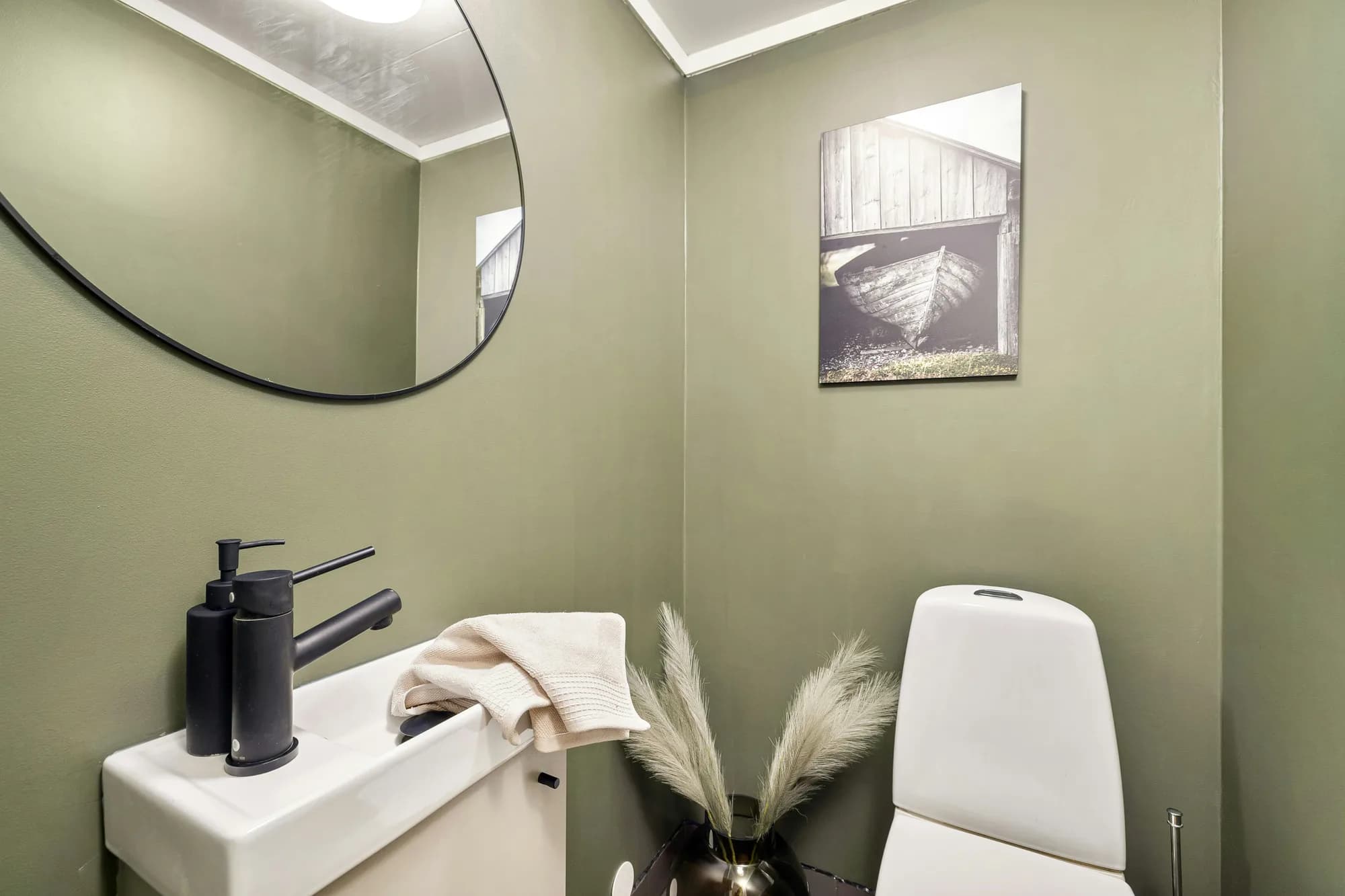 Groen geschilderde toiletruimte met een smal fonteintje, een ronde spiegel en een ingelijste foto aan de wand
